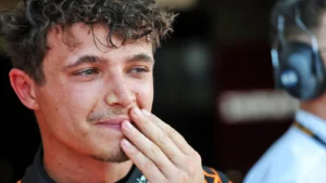 Close-up of a young man with curly dark hair touching his lips with his fingers, wearing a racing suit in a pit area.