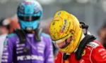 Racing drivers in helmets and fire suits, foreground driver in red with a yellow helmet, background driver in purple at the track paddock
