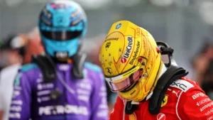 Racing drivers in helmets and fire suits, foreground driver in red with a yellow helmet, background driver in purple at the track paddock