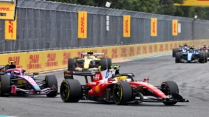 Red Formula 1 car leads a pack on a race track, with DHL banners along the barrier walls behind.