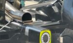 Close-up of a race car's carbon fiber body with a circular exhaust outlet and a yellow-framed wheel opening nearby.