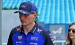 Male race car driver in a blue racing suit and cap, standing at a track with a woman wearing sunglasses in the background.