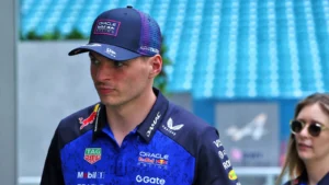 Male race car driver in a blue racing suit and cap, standing at a track with a woman wearing sunglasses in the background.