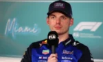 F1 driver in a blue and black racing suit speaks at a press conference, holding a FIA microphone with a Miami backdrop.