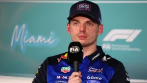 F1 driver in a blue and black racing suit speaks at a press conference, holding a FIA microphone with a Miami backdrop.