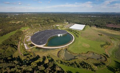 Aerial view of a park featuring a large circular building by a water body and a white rectangular structure among trees and fields, under a blue sky.