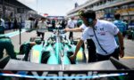 Pit stop scene with teal race car being serviced by crew in white uniforms and teal headsets in the pit lane