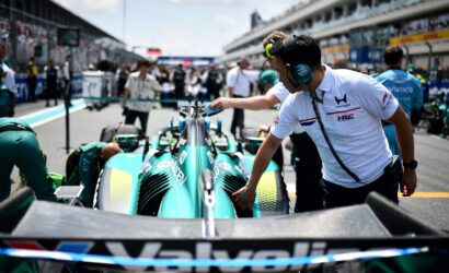 Pit stop scene with teal race car being serviced by crew in white uniforms and teal headsets in the pit lane