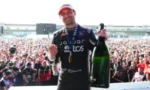 Male cyclist on a podium holding a large champagne bottle, medal around his neck, crowd cheering behind him.