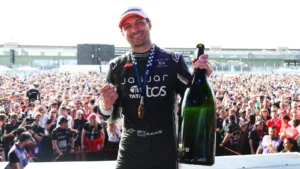 Male cyclist on a podium holding a large champagne bottle, medal around his neck, crowd cheering behind him.