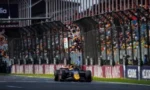 Formula 1 car racing down a track with crowded grandstands and safety fencing in the background.