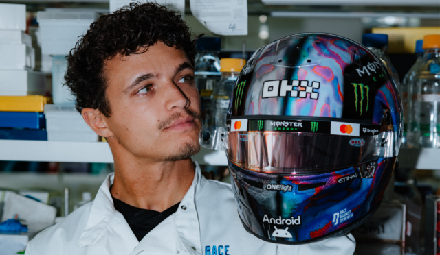 Man in a white lab coat standing next to a glossy, colorful racing helmet with Monster Energy logos.