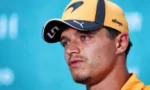Close-up of a male athlete wearing an orange and black cap, speaking at a press event with a blue background.