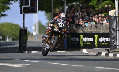 Moto racer in full gear on an orange-black bike, leaning through a turn with wheel off the ground, number 14, spectators behind barriers and Monster Energy banners in the background.