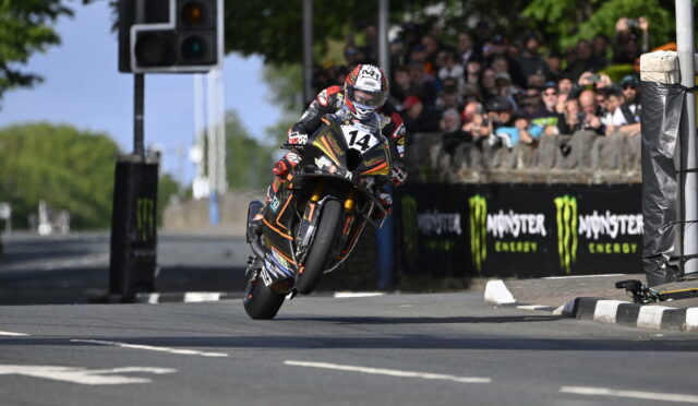 Moto racer in full gear on an orange-black bike, leaning through a turn with wheel off the ground, number 14, spectators behind barriers and Monster Energy banners in the background.