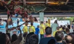 Crowd watching a live outdoor stage performance; several people in bright yellow shirts on stage under a decorated pavilion.