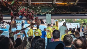 Crowd watching a live outdoor stage performance; several people in bright yellow shirts on stage under a decorated pavilion.