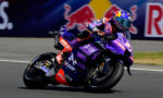 MotoGP racer in purple Yamaha livery leaning into a turn on a racing track, with a Red Bull backdrop in the background.