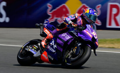 MotoGP racer in purple Yamaha livery leaning into a turn on a racing track, with a Red Bull backdrop in the background.