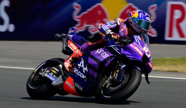 MotoGP racer in purple Yamaha livery leaning into a turn on a racing track, with a Red Bull backdrop in the background.