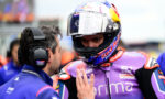 Racing rider in a purple suit and helmet talks with a crew member wearing a headset on a pit lane.