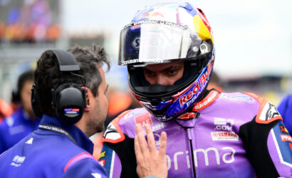 Racing rider in a purple suit and helmet talks with a crew member wearing a headset on a pit lane.