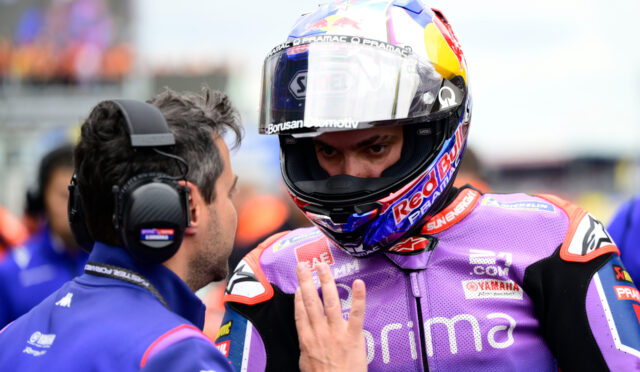 Racing rider in a purple suit and helmet talks with a crew member wearing a headset on a pit lane.