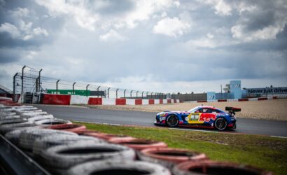 Race cars speed around a track with stacked tires in the foreground and a cloudy sky above the circuit fence and barriers.