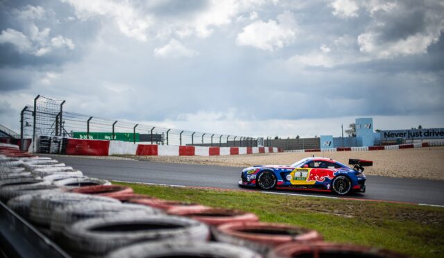 Race cars speed around a track with stacked tires in the foreground and a cloudy sky above the circuit fence and barriers.