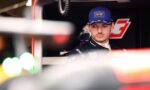 Portrait of a race car driver in a blue Oracle Red Bull cap, in a pit/garage setting with sponsor logos on the suit above.