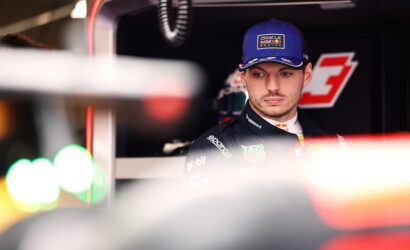 Portrait of a race car driver in a blue Oracle Red Bull cap, in a pit/garage setting with sponsor logos on the suit above.