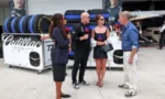 Five people are interviewed in a race pit, with stacked tires and a Cadillac-branded container in the background.