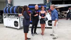 Five people are interviewed in a race pit, with stacked tires and a Cadillac-branded container in the background.