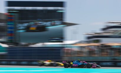 Blurry image of a Formula 1 car racing on a turquoise track, with a grandstand in the background.