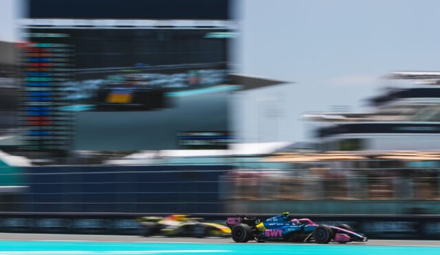 Blurry image of a Formula 1 car racing on a turquoise track, with a grandstand in the background.