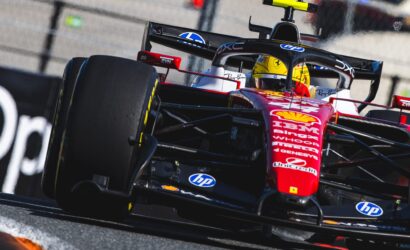 Red Formula 1 car with a yellow helmeted driver speeding along the track, close to the wall and barriers, sponsor logos visible.