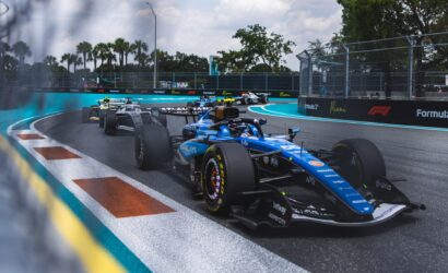 Formula 1 cars race around a tight Miami hairpin, blue track with turquoise runoff and barrier walls, palm trees in the background.