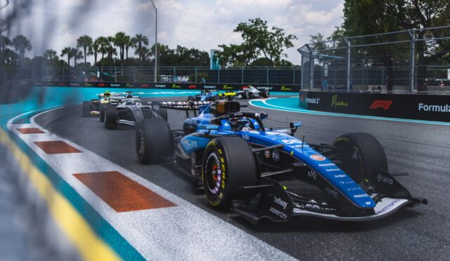 Formula 1 cars race around a tight Miami hairpin, blue track with turquoise runoff and barrier walls, palm trees in the background.