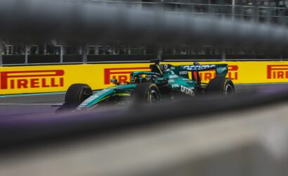 Green Formula 1 car speeding along a track with yellow Pirelli barriers in the background, viewed through a blurred foreground barrier and fence.