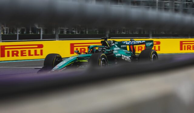 Green Formula 1 car speeding along a track with yellow Pirelli barriers in the background, viewed through a blurred foreground barrier and fence.