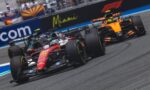 Red Ferrari Formula 1 car in the foreground on a race track, with an orange McLaren close behind and blurred spectators and banners in the background.