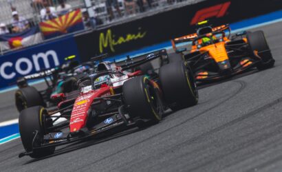 Race-pace scene with multiple Formula 1 cars speeding on the track and blurred grandstands in the background, including an orange McLaren ahead of a red Ferrari.