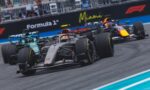 Formula 1 race cars speed along a circuit, with Miami branding on the wall and spectators in the stands behind them.