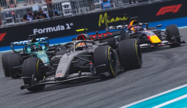 Formula 1 race cars speed along a circuit, with Miami branding on the wall and spectators in the stands behind them.
