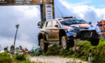 Rally car mid-air over a dirt hill during a race, with sponsor banners and a Red Bull sign in the background.