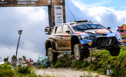Rally car mid-air over a dirt hill during a race, with sponsor banners and a Red Bull sign in the background.