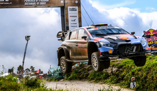 Rally car mid-air over a dirt hill during a race, with sponsor banners and a Red Bull sign in the background.