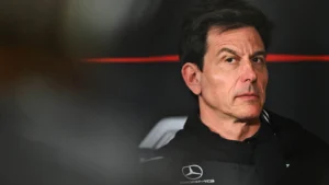 Man in a black racing jacket with a visible Mercedes emblem, looking to the right against a dark background.