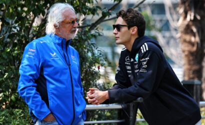 Two men are in a conversation outdoors, one in a bright blue jacket and the other in a dark athletic hoodie, near greenery.