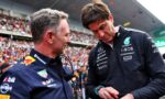 Two men in dark blue racing team jackets talk in a crowded stadium, one in profile facing left and the other looking down, with fans in the background.